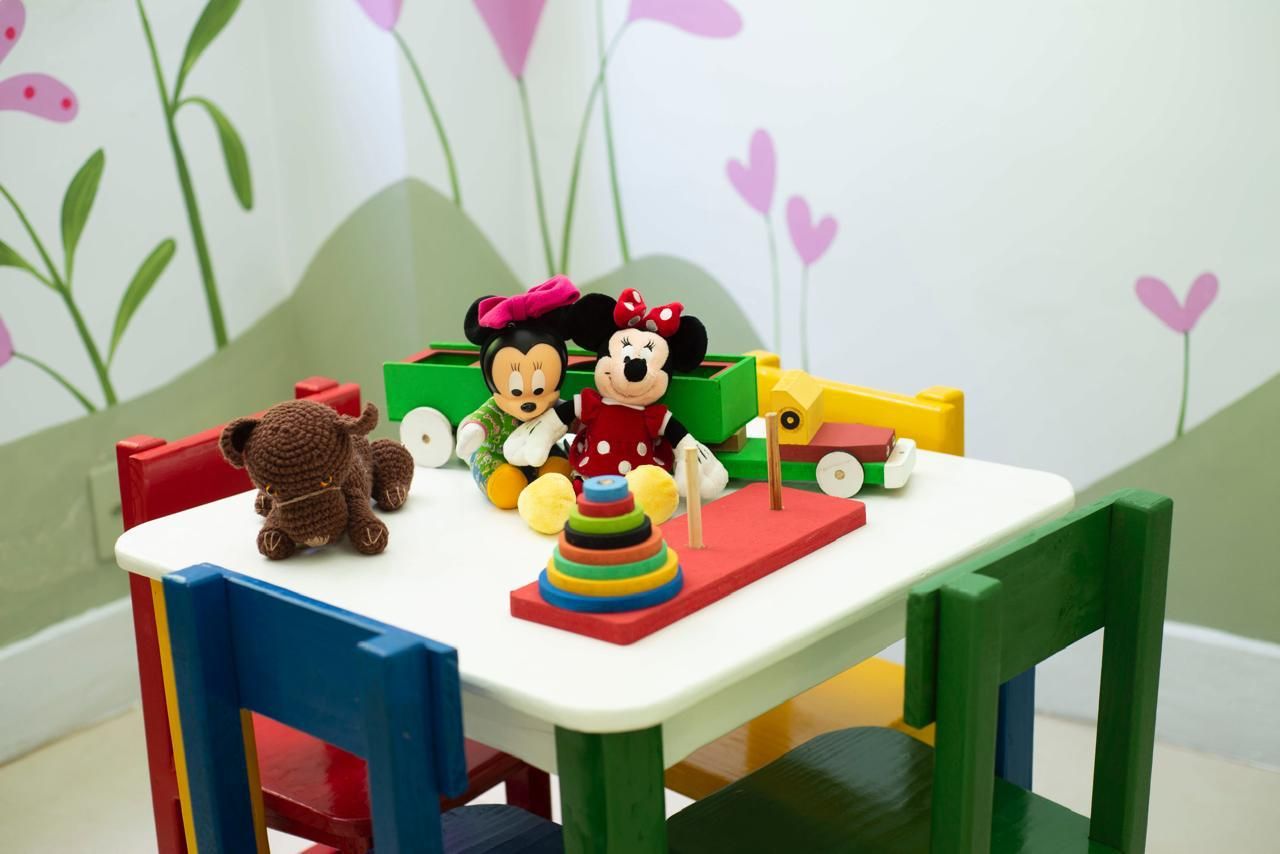 Mesinha infantil com cadeiras coloridas, bichos de pelúcia e brinquedos de madeira, diante de um mural de flores