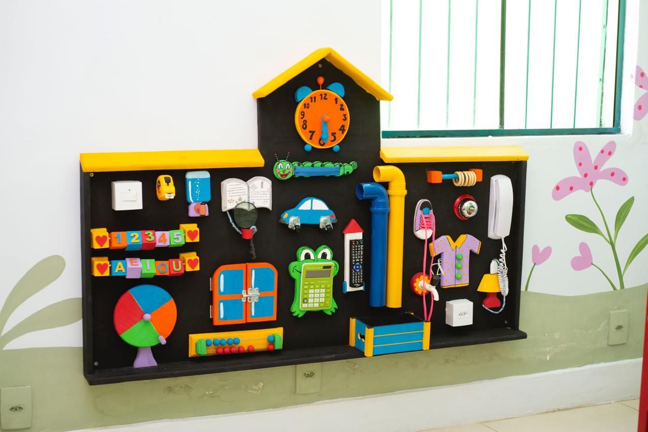 Painel de atividades em forma de casinha, com relógio, letras, números, trincos e objetos do dia a dia para as crianças manusearem enquanto esperam